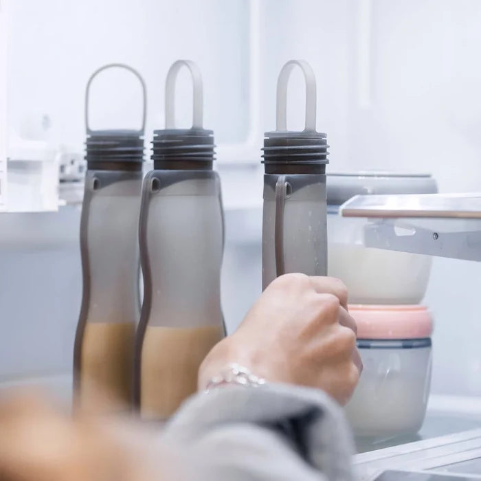 An eco-conscious parent chooses from three frosted bottles with looped handles stored in a fridge. Nearby, containers with pink and white lids stack, housing baby-feeding essentials like Haakaas Silicone Milk Storage Bag & Sippy Spout Combo Pack.