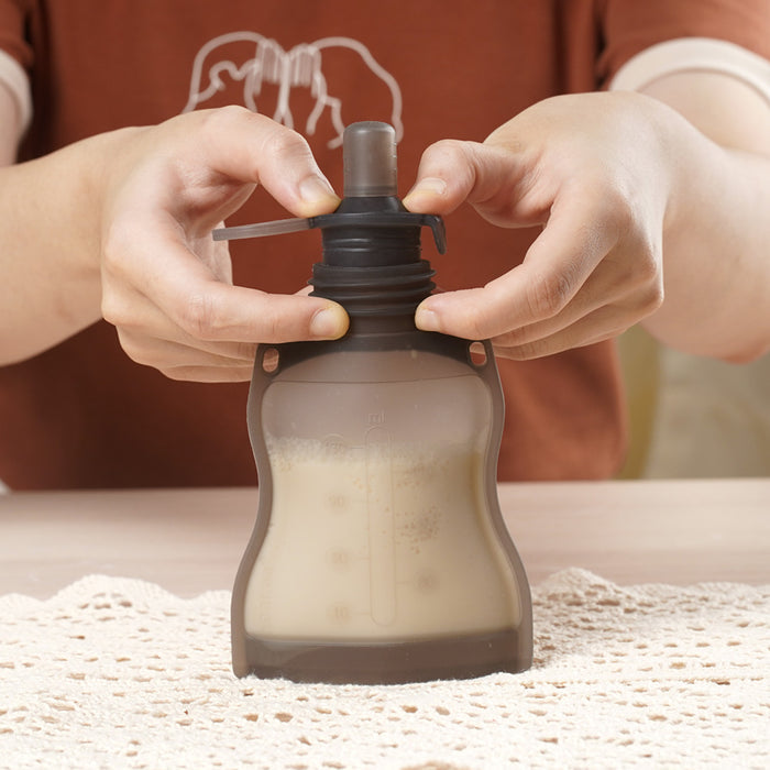 A person handles a partially filled Haakaa Silicone Milk Storage Bag with a black sippy spout, pressing it open. In the background, an eco-conscious parent in a brown shirt stands by a laced table to keep baby-feeding essentials within reach.