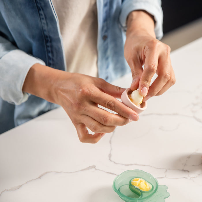 On the marble countertop, a person in a denim jacket presses a hard-boiled egg into a small gadget. Nearby, a green dish with a peeled egg suggests they use the Haakaa Silicone Freeze-N-Feed Mini Combo for teething relief.