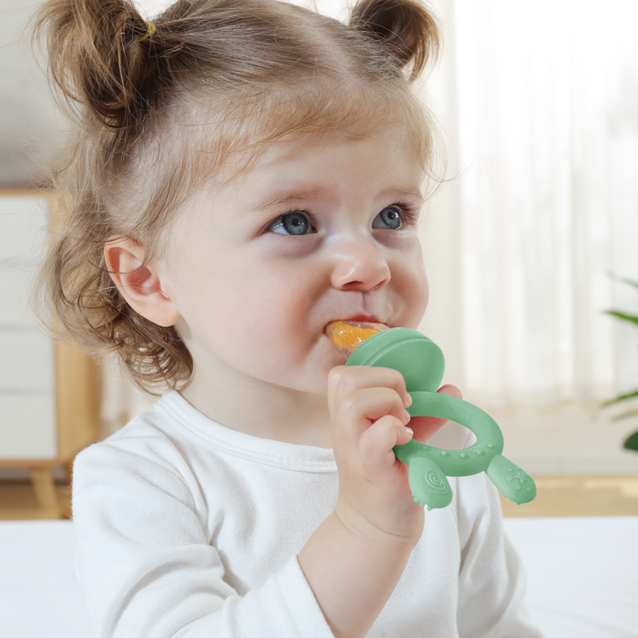A toddler with light brown pigtails sits indoors in a white top, holding a green Haakaa Silicone Freeze-N-Feed Mini Combo feeder. She finds teething relief as she chews on it, against a softly blurred background that suggests a cozy home environment.