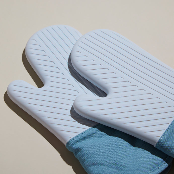 The GIR silicone oven mitts, in light blue with textured surfaces and fabric cuffs, offer excellent kitchen protection. Placed side by side on a neutral background, they beautifully blend contrasting textures while ensuring reliable heat resistance.