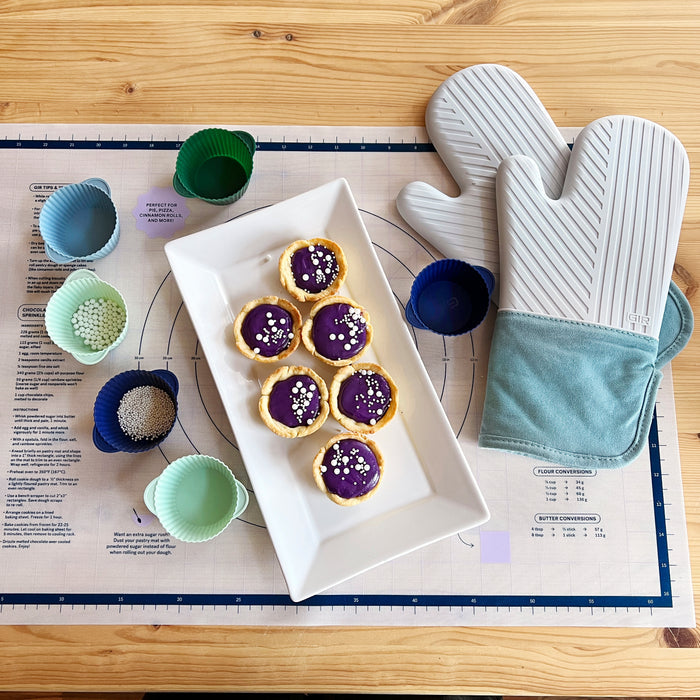 On a wooden table are six purple tarts on a rectangular plate, colorful cupcake liners with sprinkles, and a pastry mat. Nearby is a pair of heat-resistant, BPA-free silicone oven mitts by GIR.