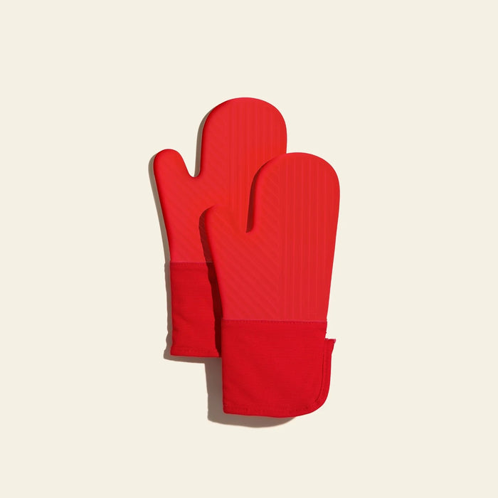 Two bright red GIR Silicone Oven Mitts, featuring heat-resistant, BPA-free silicone with textured surfaces and fabric cuffs, rest against a light beige background, slightly overlapping.