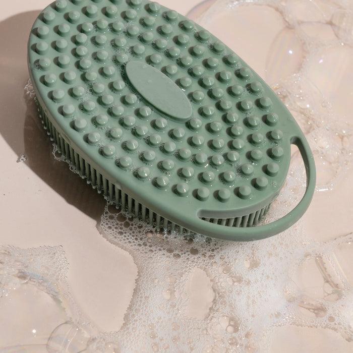 The EcoTools Silicone Scrubber, an eco-friendly green oval scrubber with raised bumps and a handle, sits on a light surface surrounded by soap bubbles.