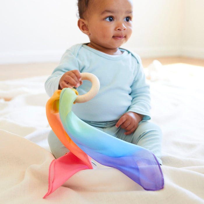 A baby sits on a soft surface holding a Sarahs Silks Silk and FSC-Certified Wood Baby Teether adorned with vibrant scarves in pink, orange, yellow, green, and blue. Made with eco-friendly dyes, it matches their light blue outfit as they gaze sideways in the bright, softly lit background.