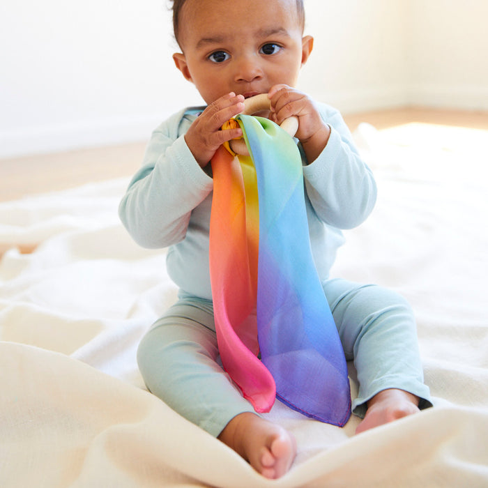 A baby in a light blue onesie sits on a white blanket, holding a vibrant rainbow scarf and Sarahs Silks Silk and FSC-Certified Wood Baby Teether. The baby looks directly at the camera as natural light fills the room.