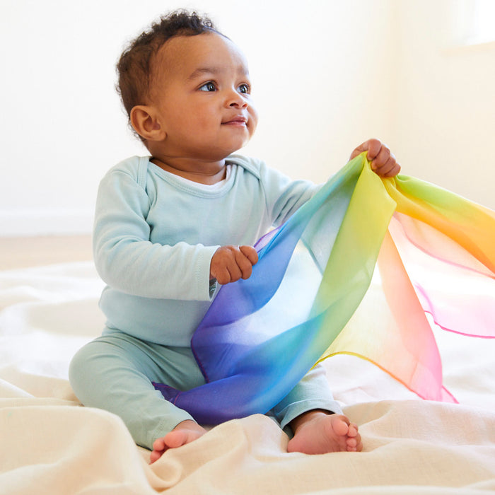 A baby in a light blue outfit sits on a soft blanket, holding Sarahs Silks vibrant Silk and FSC-Certified Wood Baby Teether, made with eco-friendly dyes. The room is bright and softly lit, creating a warm, playful atmosphere.