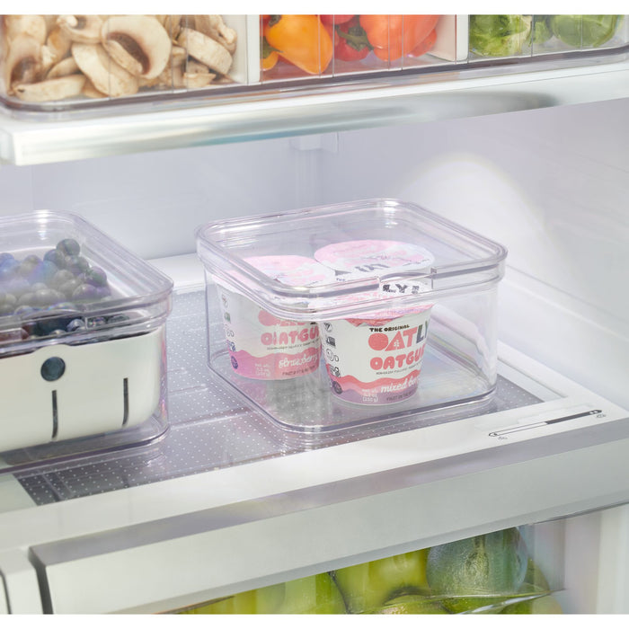 An iDesign Single Fridge Bin made with recycled plastic holds two tubs of Oatly Oatgurt Mixed Berry beside a container of blueberries, while neatly stored assorted vegetables sit above in clear, BPA-free bins.