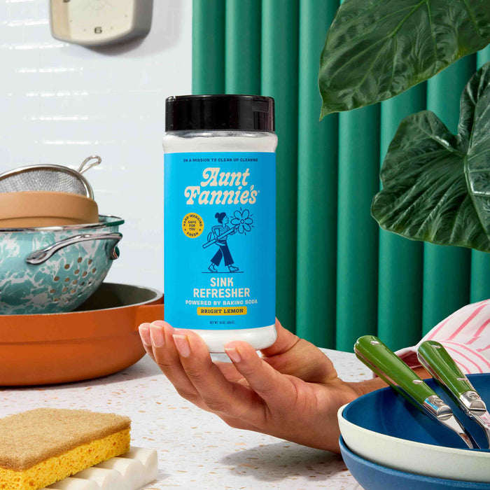 A hand holds a container of Aunt Fannies Sink Refresher over a kitchen counter with dishes, utensils, a sponge, and green plants in the background.