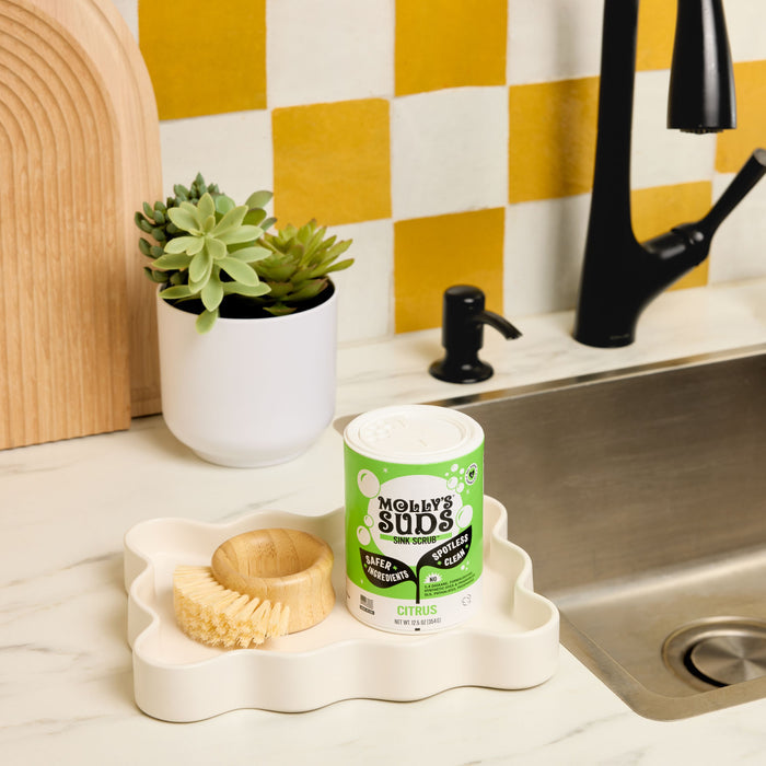 A canister of Molly’s Suds Sink SCRUB™ Sink Cleaner, a round wooden brush, and a small potted succulent sit on a wavy white tray by a kitchen sink with a black faucet, against a yellow and white checkerboard backsplash.