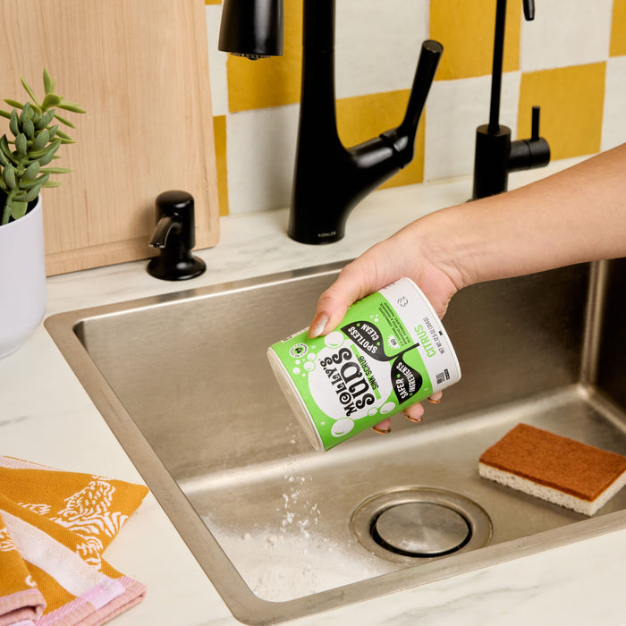 A hand pours Molly's Suds Sink SCRUB™ Sink Cleaner into a kitchen sink. Nearby are a brown sponge, soap dispenser, small potted plant, and folded yellow and pink towel—everything for a gentle scrub.