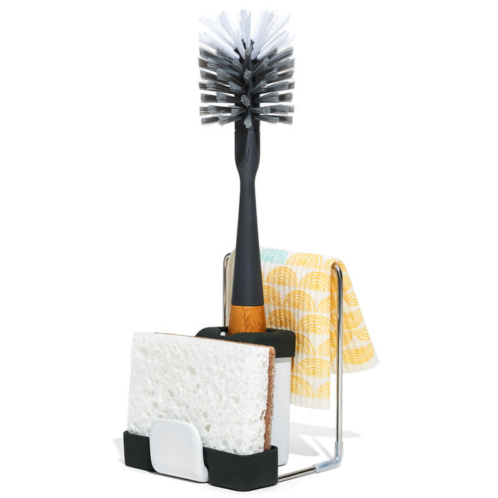 The Grove Co. Sink-Side Caddy, crafted from metal with elegant black accents, holds a dish brush with black bristles, a white and brown sponge, and a folded dish towel featuring a yellow and blue circular pattern. Its plastic-free and durable.