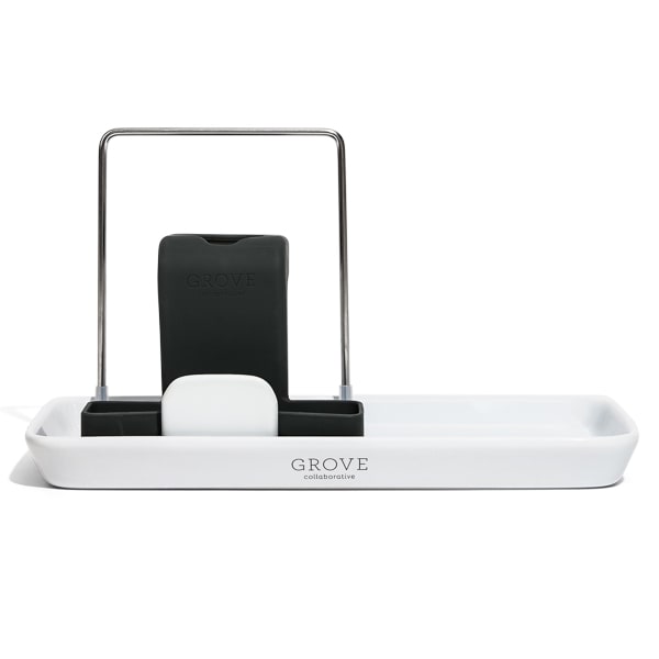 The Grove Co. Sink-Side Caddy features a white rectangular ceramic tray with GROVE inscribed, cradling a black scrubber and soap bar on a metal frame, making it a durable, plastic-free, minimalistic organizer for your sink area.