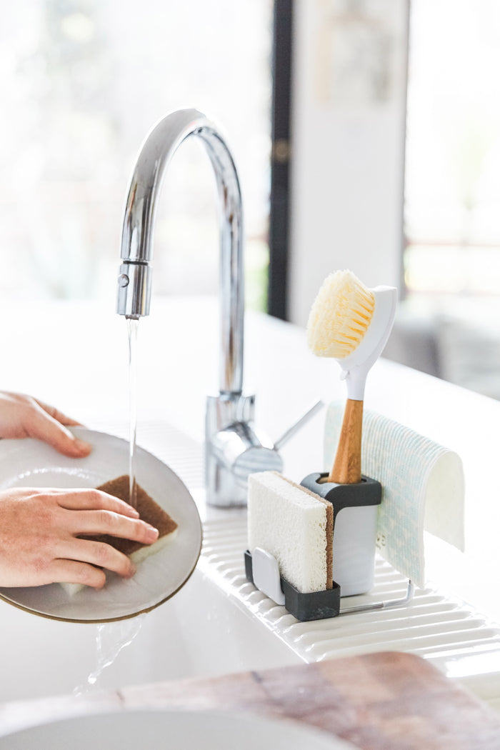 In a softly blurred kitchen, a person washes a plate while the Grove Co. Sink-Side Caddy neatly holds a dish brush, sponge, and scrubber beside the sink. A dishcloth hangs over the caddy.