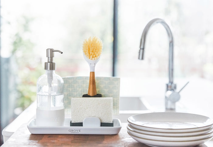 A kitchen sink with a silver faucet includes Grove Co.s plastic-free Sink-Side Caddy, which holds a soap dispenser, dish brush, sponge, and patterned cloth. Nearby, gleaming white plates are illuminated by bright natural light.
