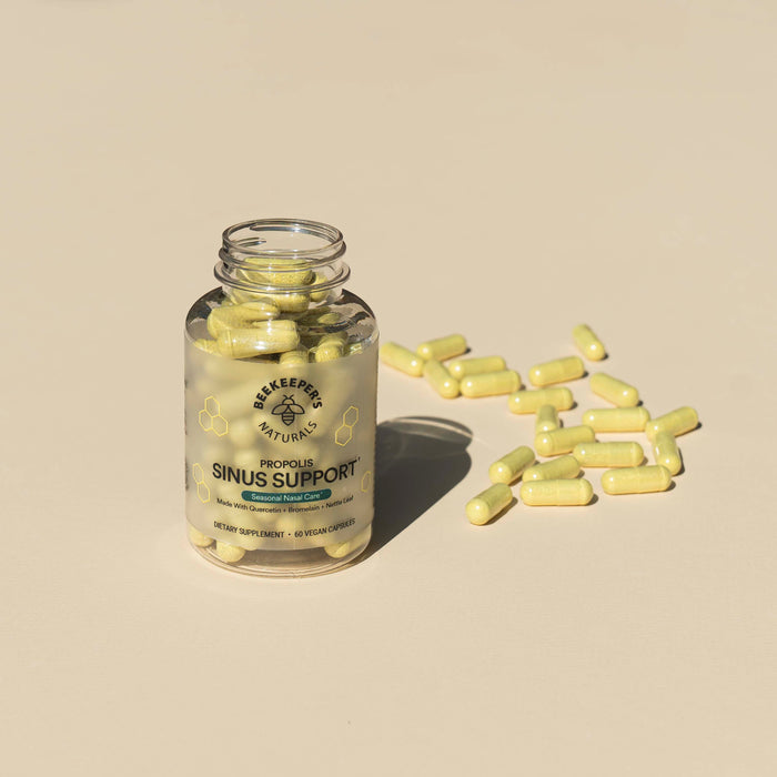 A clear jar labeled Sinus Support by Beekeepers Naturals contains yellow capsules for sinus and immune aid. Several capsules are scattered on a light beige surface next to the jar, whose label with a small bee logo highlights its role in maintaining healthy histamine levels.