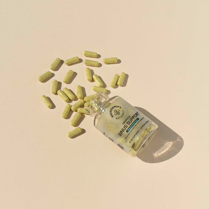 A clear bottle labeled Sinus Support by Beekeepers Naturals is on a light beige surface with green capsules spilling out. These uniform capsules provide sinus and immune support, casually arranged around for easy access.