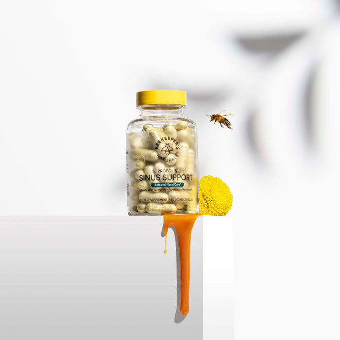A Beekeepers Naturals Sinus Support bottle with a yellow cap rests on a surface. Honey spills from its base, pooling by a yellow flower. Nearby, a bee hovers against a white background, symbolizing natural wellness.