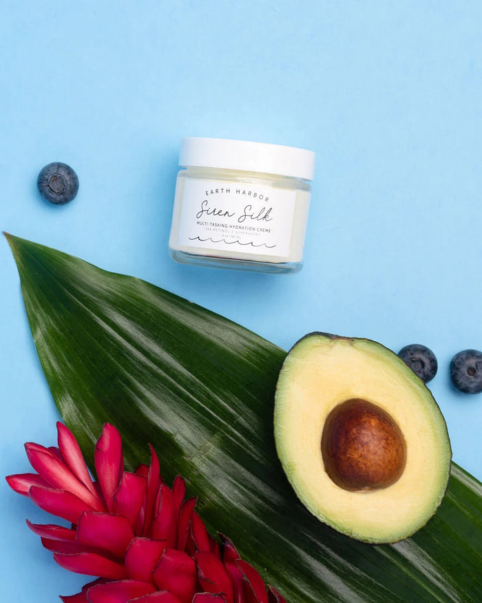 A jar of Earth Harbors SIREN SILK Bio-Retinol Hydration Cream sits on a blue background among half an avocado, a red tropical flower, a lush green leaf, and three blueberries—creating the ideal setting for this versatile treatment cream.