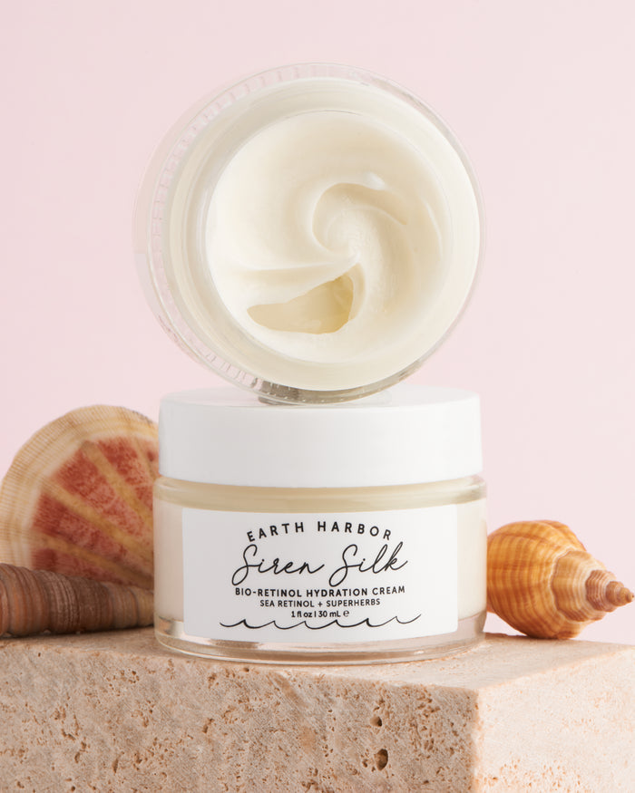 An open jar of SIREN SILK Bio-Retinol Hydration Cream by Earth Harbor displays its creamy texture. It sits on a stone surface with seashells and a soft pink backdrop, capturing the essence of elegant anti-aging skincare.