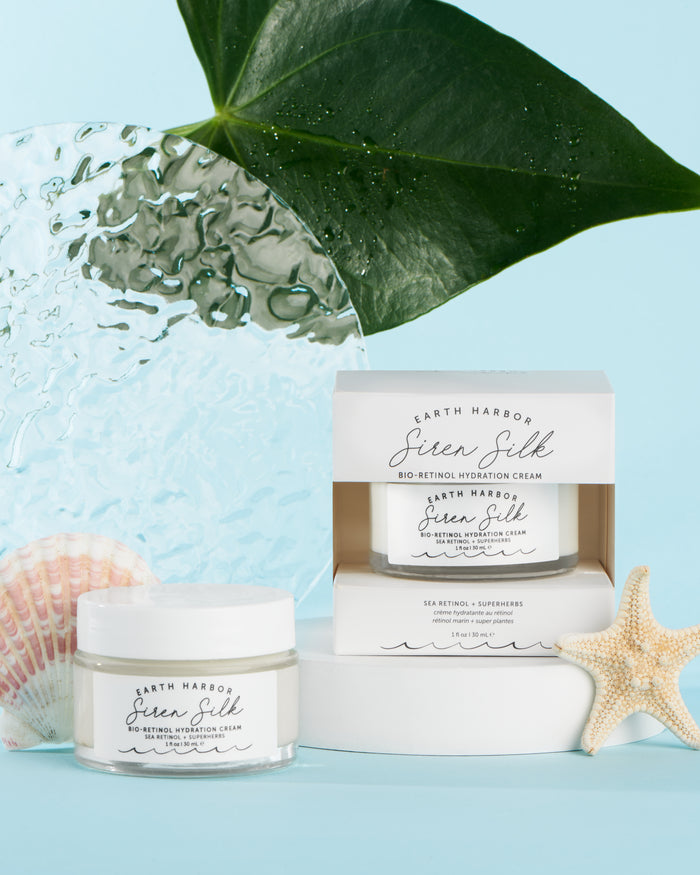 Displayed against a light blue background are a jar and box of Earth Harbors SIREN SILK Bio-Retinol Hydration Cream, an anti-aging multitasking treatment. They are accompanied by a starfish, seashell, textured glass panel, and large leaf.