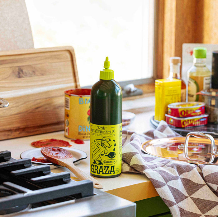 A kitchen counter features a bottle of Grazas Sizzle Extra Virgin Olive Oil for cooking, sitting with essentials like canned fish, a spice jar, and tomato sauce. A pot waits on the stove as bright natural light streams in through the window.