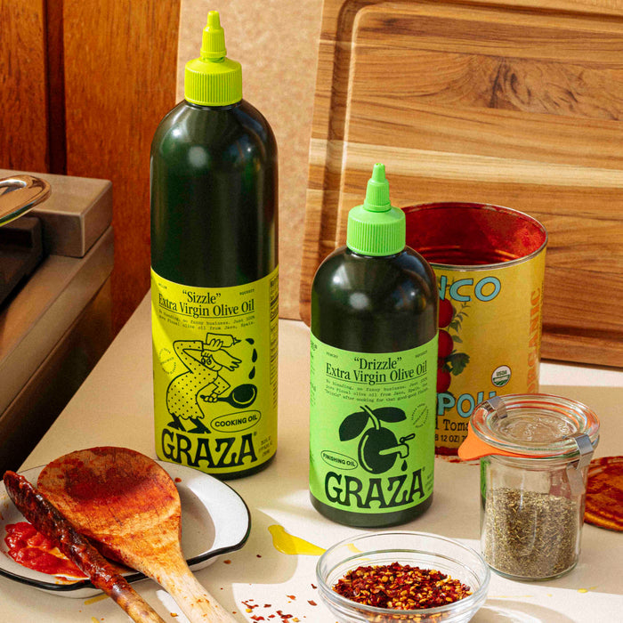 Two bottles of Graza extra virgin olive oil, one labeled Sizzle Extra Virgin Olive Oil - For Cooking, sit on a kitchen counter with green caps alongside an open can of tomatoes, a spice jar, a wooden spoon with sauce, red pepper flakes, and spilled mellow-flavored olive oil.