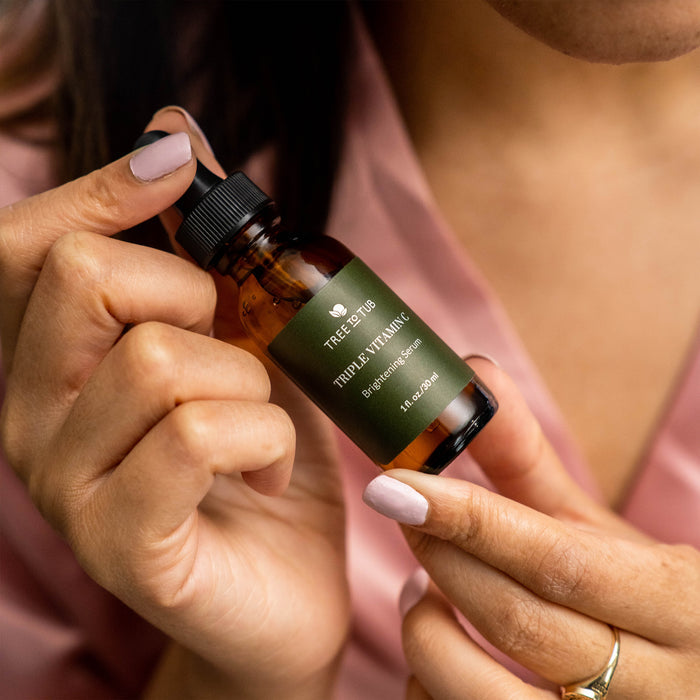 A person with light purple nails holds a brown bottle of Tree To Tubs Skin Brightening Triple Action Vitamin C Serum for Face. The anti-aging bottle, featuring a black dropper cap and green label, is carefully adjusted by the person wearing a pink garment.