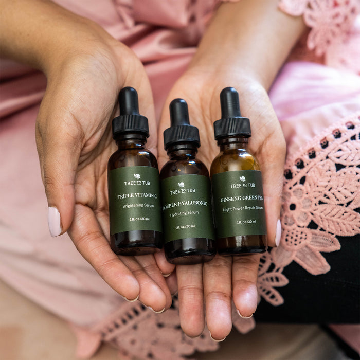 A person in a pink lace-trimmed garment holds three Tree To Tub bottles labeled: Skin Brightening Triple Action Vitamin C Serum for Face, Pure Hyaluronic Hydrating Serum, and Ginseng Green Tea Night Power Repair Serum.