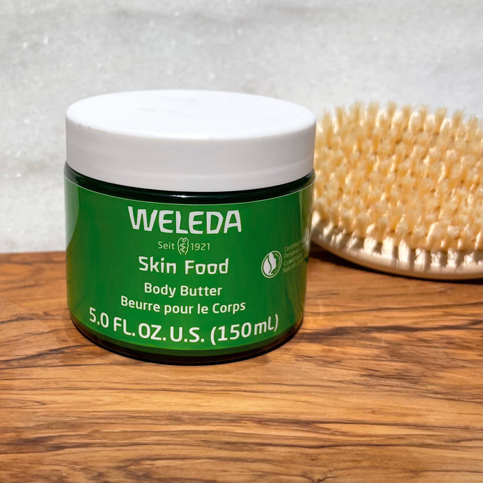 A 5.0 fl. oz. (150 ml) green jar of Weleda Skin Food Body Butter, packed with shea and cocoa seed butters for non-greasy hydration, rests on a wooden surface with a natural bristle body brush in the background, providing a complete skincare experience.