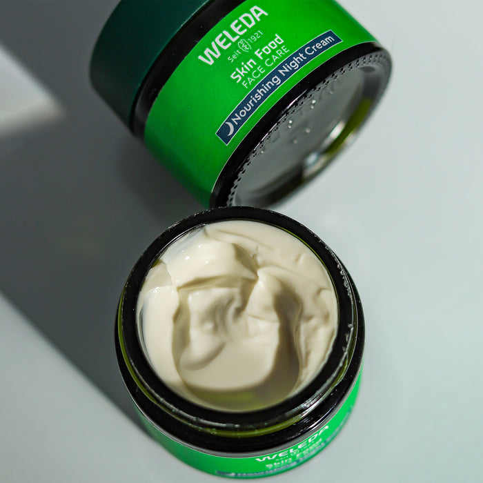 A green jar of Weleda Skin Food Face Care Nourishing Night Cream with white butter face cream sits on a light surface. The lid is partially visible beside it, with shadows providing a subtle contrast, enhancing its promise to nourish skin overnight.