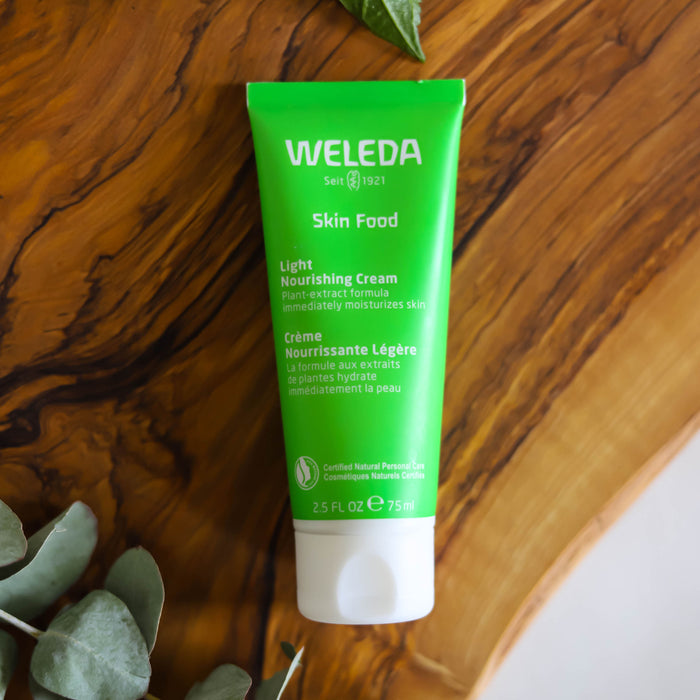 A 75 ml tube of Weleda Skin Food Light Nourishing Cream sits on wood, its green packaging emphasizing a plant-extract formula for deep moisturizing. Green leaves in the corner accentuate the natural theme.