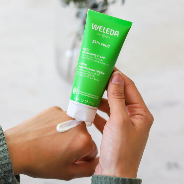 A person squeezes Weledas Skin Food Light Nourishing Cream from a green tube onto the back of their hand while another hand holds the cream, set against a blurred background.