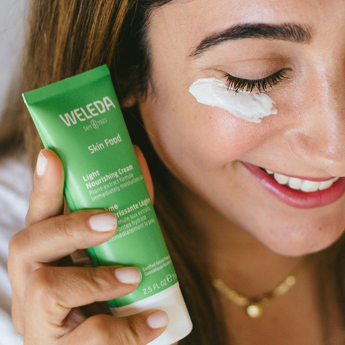 A person smiles while massaging Skin Food Light Nourishing Cream by Weleda onto their face. They hold the green tube near their cheek with a visible dab of moisturizer by their eye.