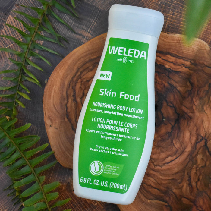 A 6.8 fl oz (200 ml) bottle of Weledas Skin Food Nourishing Body Lotion sits on wood, surrounded by green leaves. It is enriched with plant extracts like viola tricolor and offers Intensive, long-lasting nourishment for dry to very dry skin.