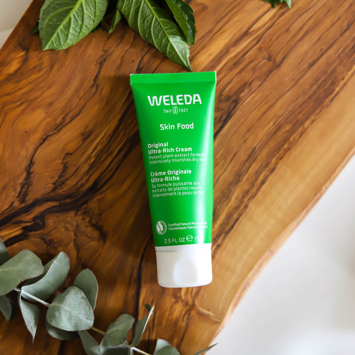 A vibrant green tube of Weledas Skin Food Original Ultra-Rich Cream moisturizer rests on a wooden surface, with its bilingual English and French label surrounded by green leaves, enhancing the natural, organic atmosphere.