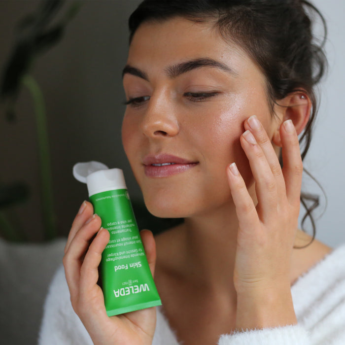 A person with dark hair in a white top gently applies Weledas Skin Food Original Ultra-Rich Cream to their face. They appear serene, standing in a softly lit room as the moisturizer from the green tube nourishes their skin.