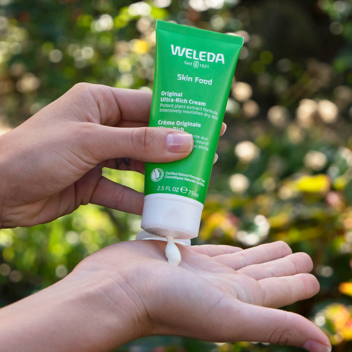 A person squeezes Weleda Skin Food Original Ultra-Rich Cream from a 2.5 fl oz (75 ml) green tube onto their palm, with blurred lush greenery in the background and visible certification logos on the packaging.