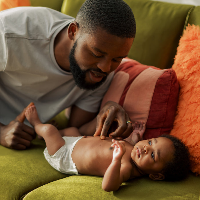 A man smiles affectionately at a baby lying on a green sofa with orange and red cushions. The baby, wearing DYPER Skin Happy Baby Diapers—gentle, high-absorbency protection with certified bamboo layers—looks up at him.