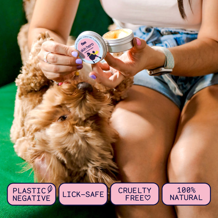 A person sitting on a green couch, holding a small tin of dog balm labeled "skin hero" while a fluffy brown dog looks at it. The text at the bottom reads "Plastic Negative," "Lick-Safe," "Cruelty Free," and "100% Natural.