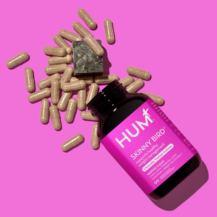 A partially open HUM Nutrition Skinny Bird bottle, with capsules spilling onto a bright pink backdrop, lies beside a small metallic weight. The label emphasizes Curbs Appetite & Weight Management and reducing stress eating.
