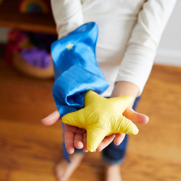 Wearing a long-sleeved white shirt, someone holds Sarahs Silks Skytail, featuring a plush yellow star and a blue cloth with small stars. The Montessori & Waldorf-inspired home has a wooden floor and blurred colorful objects in the background, perfect for open-ended play.