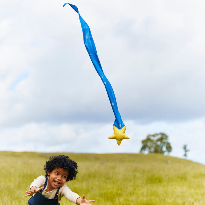 In an open grassy field, a child joyfully throws their Skytail by Sarahs Silks, featuring blue fabric with a yellow star, enhancing motor skills under the overcast sky. A distant tree adds serenity to this playful learning scene.