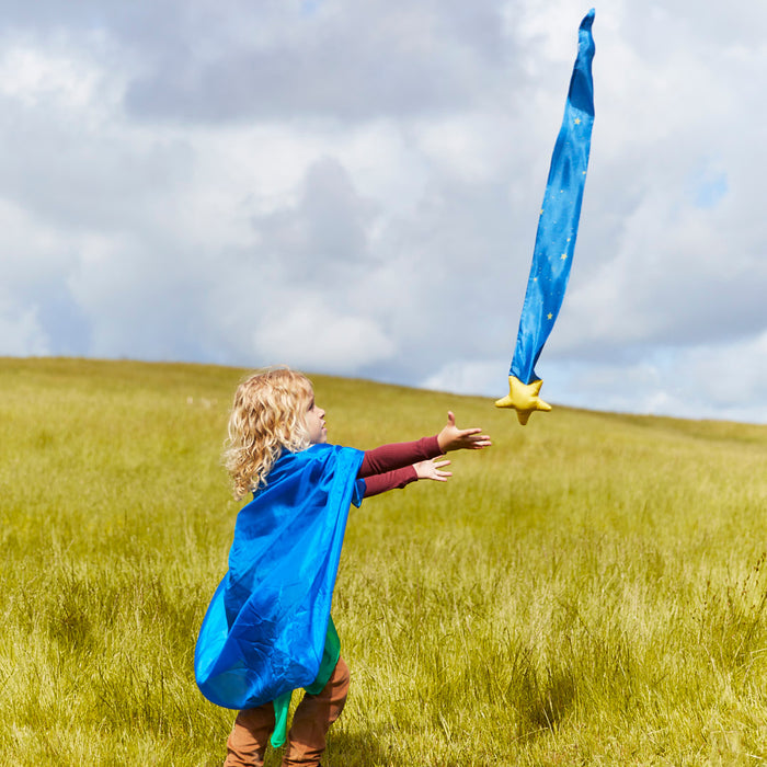 A child in a blue cape twirls in a grassy field, engaged in imaginative play with Sarahs Silks Skytail wand featuring a yellow star, embodying wonder beneath the partly cloudy sky—a moment capturing the spirit of Montessori and Waldorf homes.