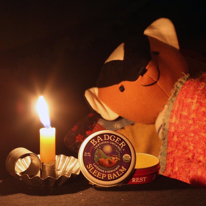 A cozy scene is softly lit by a candle, featuring Badger Balms Sleep Balm tin. Infused with calming essential oils, it complements a plush stuffed animal nestled under a quilted blanket, creating a warm atmosphere.