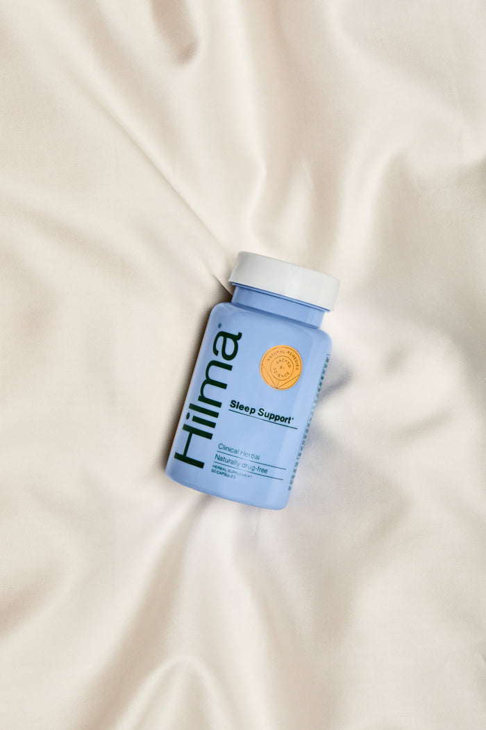 A light blue Hilma Sleep Support bottle, a natural sleep aid without melatonin, sits on cream fabric. It has a white cap and a small orange sticker near the top.