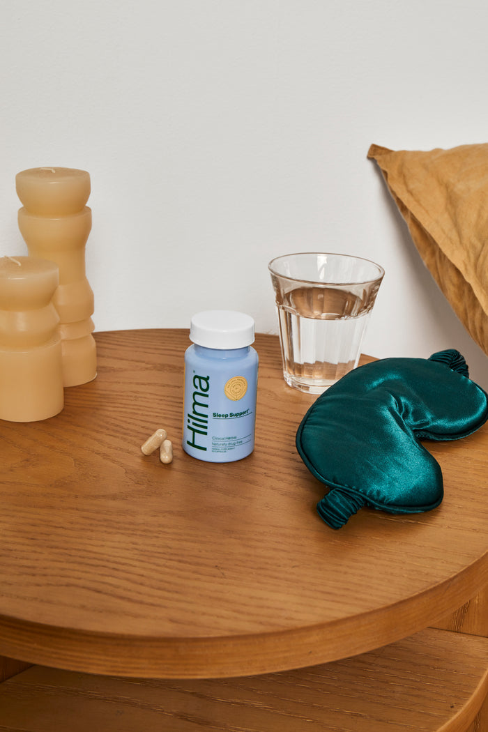 A wooden table showcases Hilmas Sleep Support, a natural, melatonin-free sleep aid, with two capsules beside a water glass. Three beige candles and a green satin sleep mask add to the tranquil scene for restful nights, with a pillow subtly in the background.