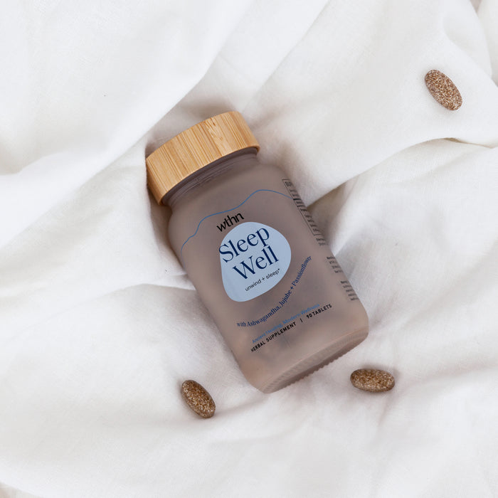 A frosted glass jar with a bamboo lid labeled Sleep Well from WTHN sits on white fabric, surrounded by three oval brown tablets. The label promotes Organic Ashwagandha and L-Theanine for natural sedation as a dietary supplement for deep sleep.