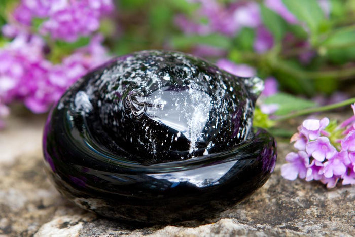 Sleeping glass cat paperweight in deep black with grey cremains under clear glass on a rocky surface with purple flowers.