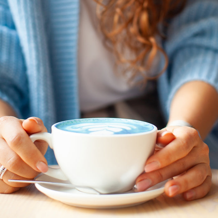 A person with curly hair and a blue sweater holds a white cup of Kuli Kuli Foods Sleeptime Organic Superfood Latte, featuring heart-shaped latte art, resting on a saucer with a spoon.